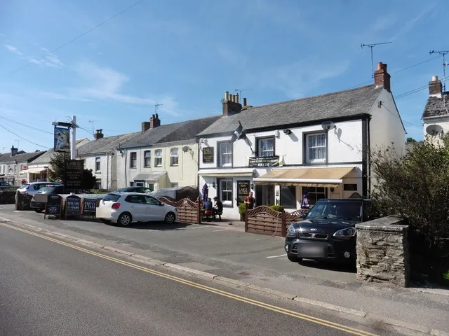 The Welcome Home Inn from across Par Green: a long white-painted pub with a grey slate roof, a cream awning over the front windows, a hanging sign on a post by the road and chalkboards along the pavement, with a fenced seating area at the front.